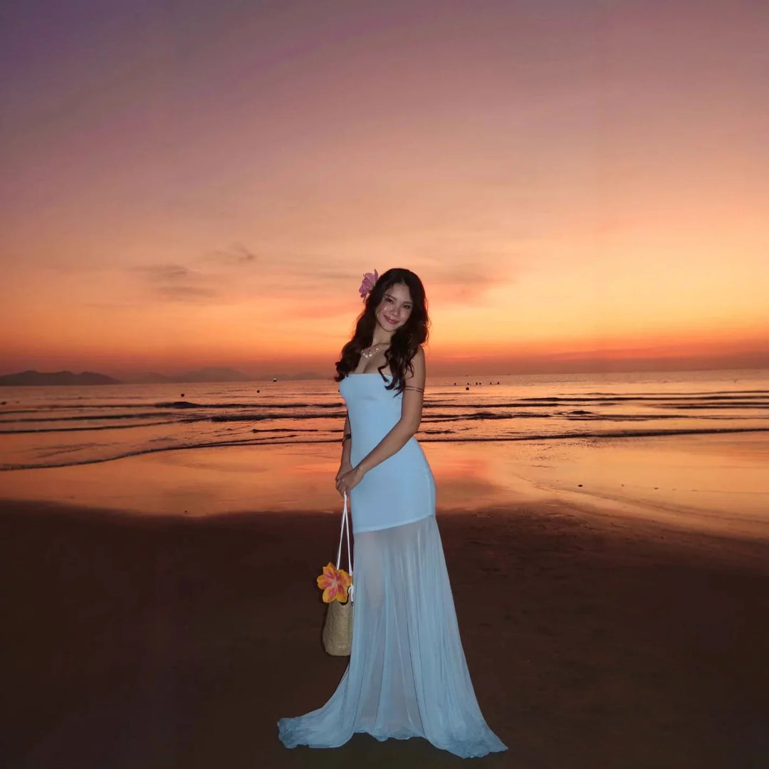 Woman in white dress with flower in hair on beach at sunset holding a bag with flowers