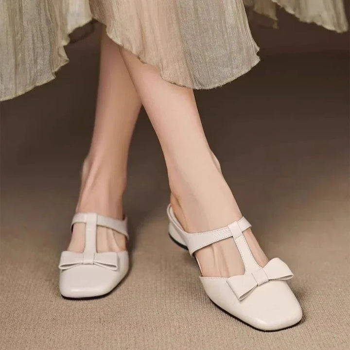 Womens cream T-strap flats with bow detail, paired with a beige pleated skirt on carpet floor