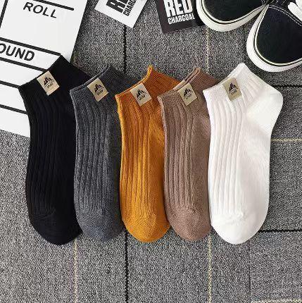 Five pairs of ribbed ankle socks in black, gray, orange, beige, and white on a gray fabric background with sneakers and film clapperboard props