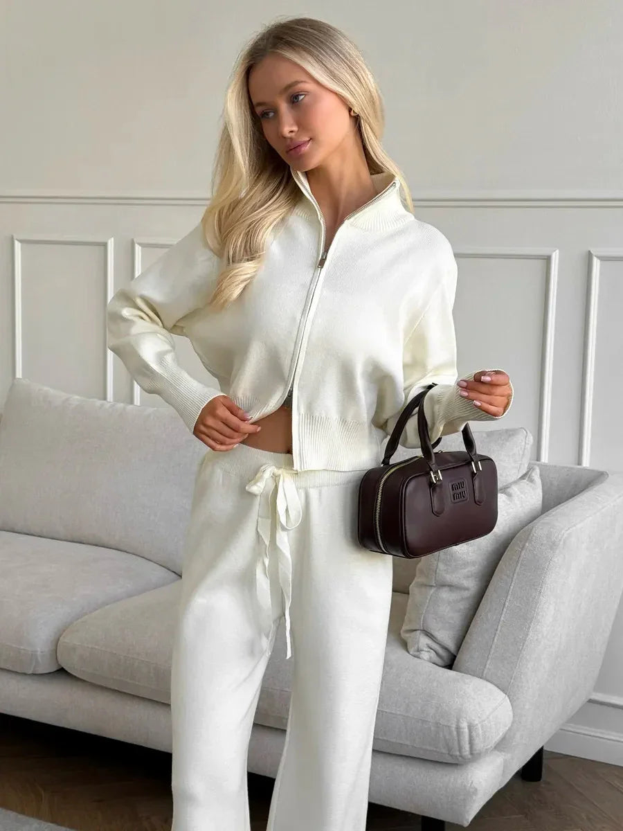 Blonde woman in white zip-up sweater and drawstring pants holding dark brown Miu Miu handbag indoors