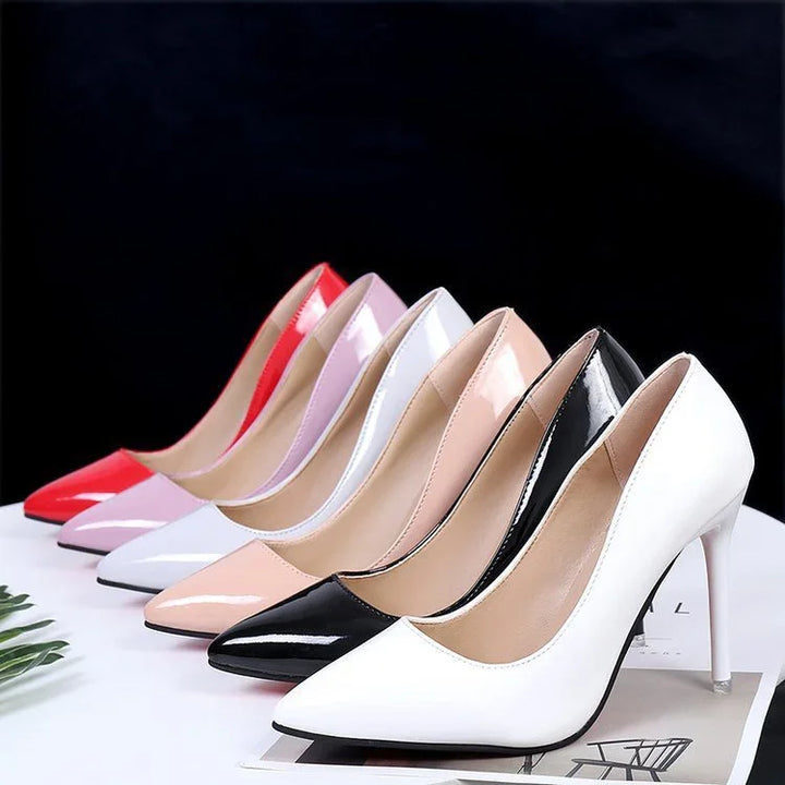 Row of glossy high-heeled pointed toe pumps in white, black, beige, white, pink, and red on a white surface