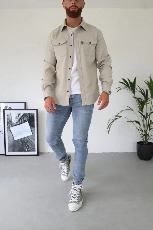 Man wearing beige button-up shirt, white tee, light blue jeans, and gray sneakers in minimal room