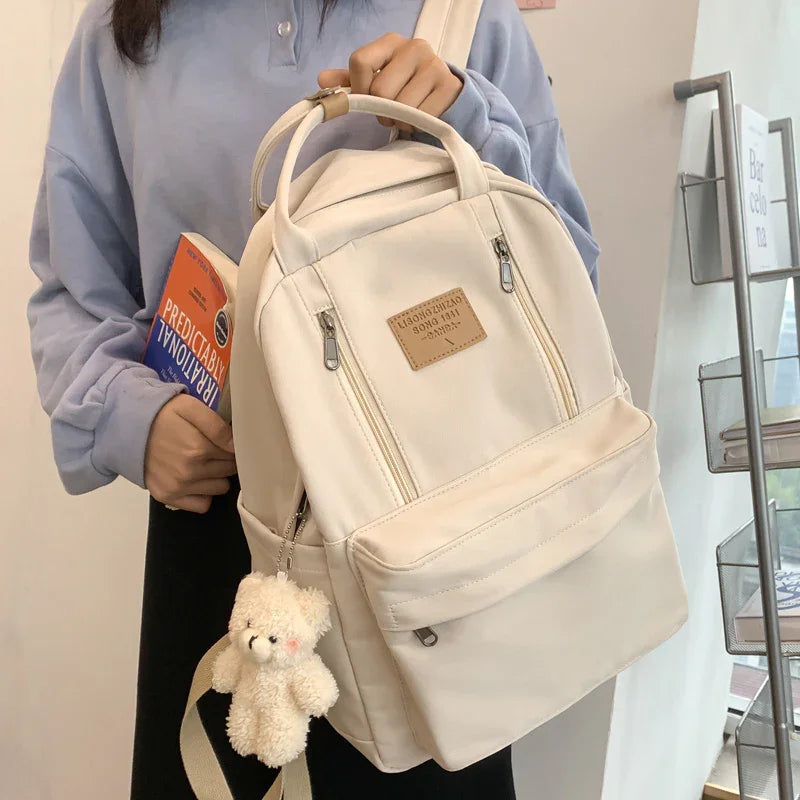 Beige backpack with front pockets and teddy bear keychain held by person in blue top holding book