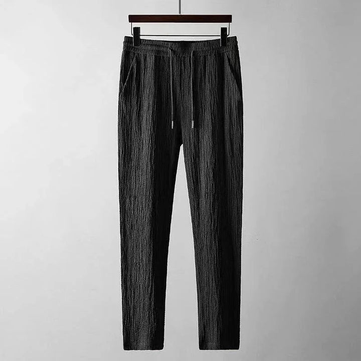 Black textured casual pants with drawstring waist hanging on wooden hanger against gray background