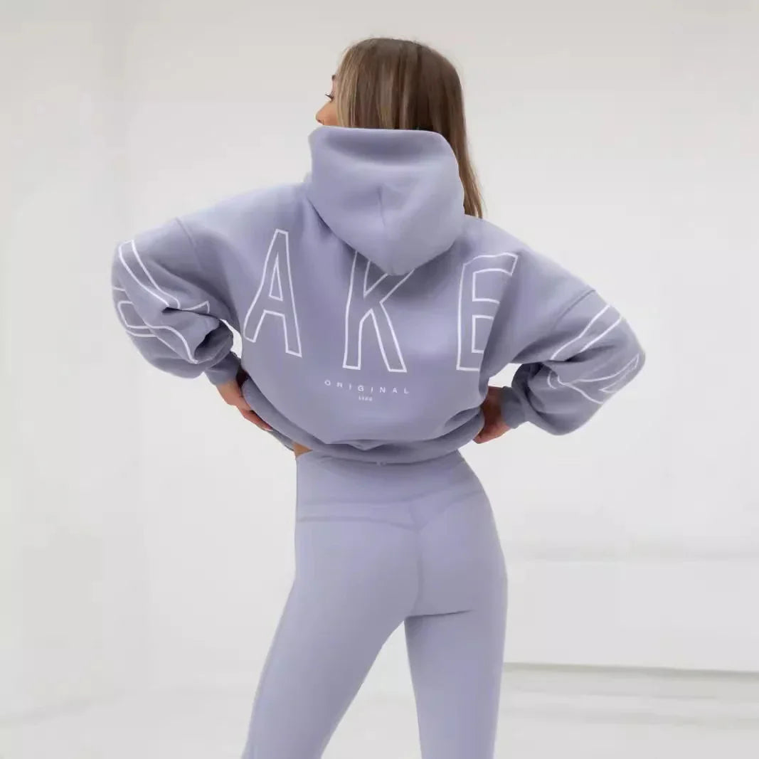 Woman in light purple hoodie and matching leggings with large text design on back, standing in bright studio