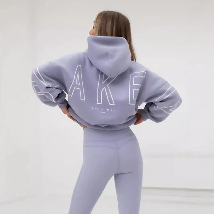 Woman in light purple hoodie and matching leggings with large text design on back, standing in bright studio
