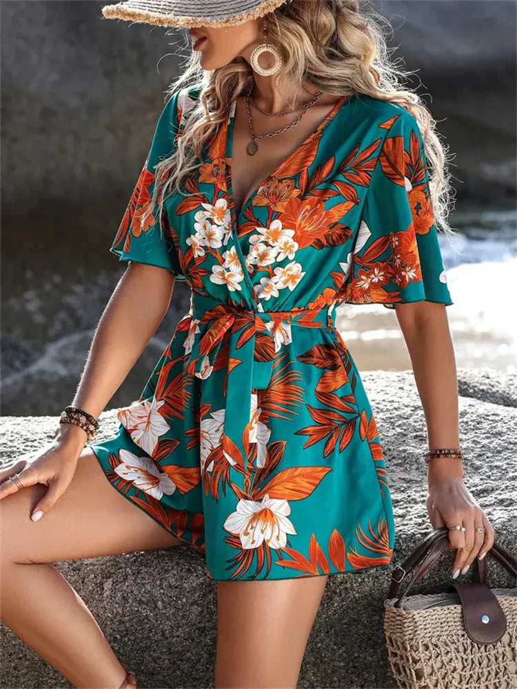 Woman wearing teal floral wrap romper with orange and white flowers, accessorized with hat and woven bag