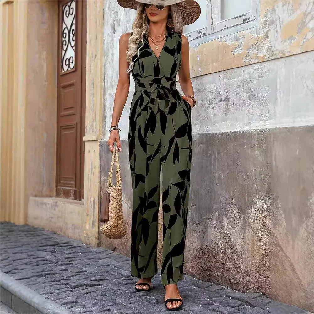 Woman in sleeveless olive green and black leaf print jumpsuit walking on cobblestone street with straw hat and woven bag