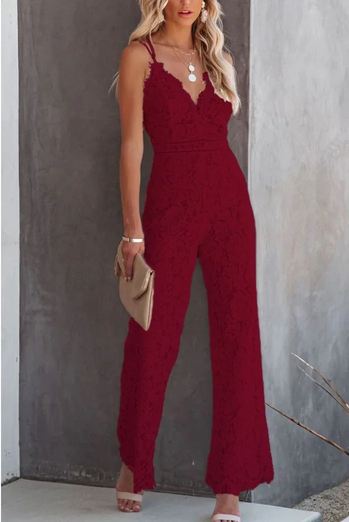 Woman wearing a burgundy lace jumpsuit with spaghetti straps holding a beige clutch against a gray concrete wall