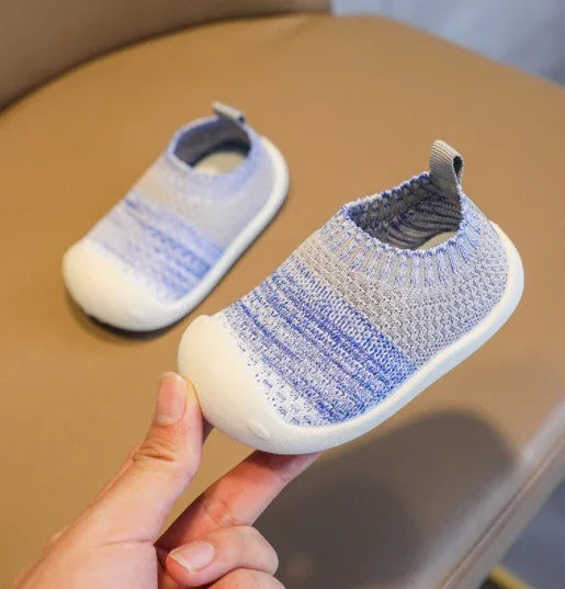 Soft knit baby shoes in gray and blue with white rubber soles held over brown couch