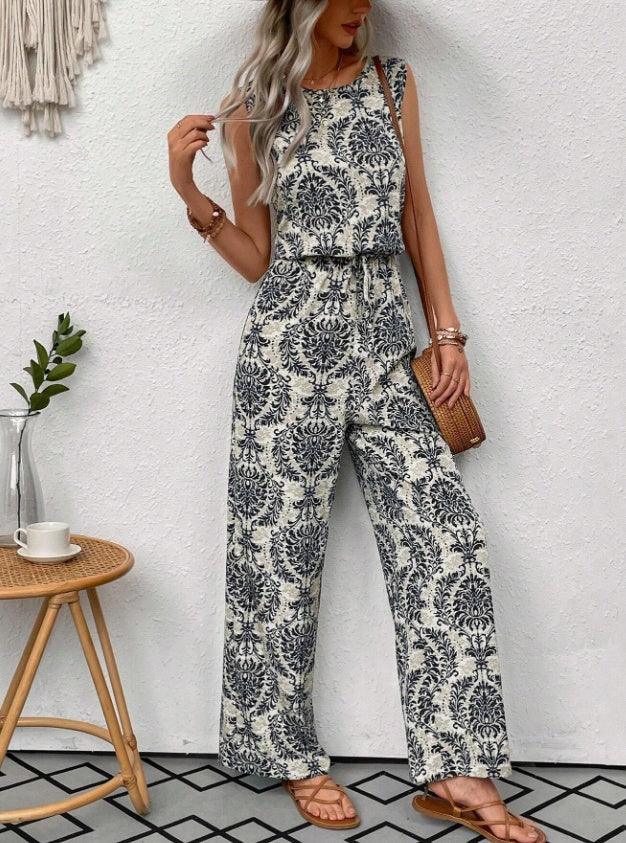 Woman wearing sleeveless wide-leg jumpsuit with black floral pattern and sandals standing against white wall