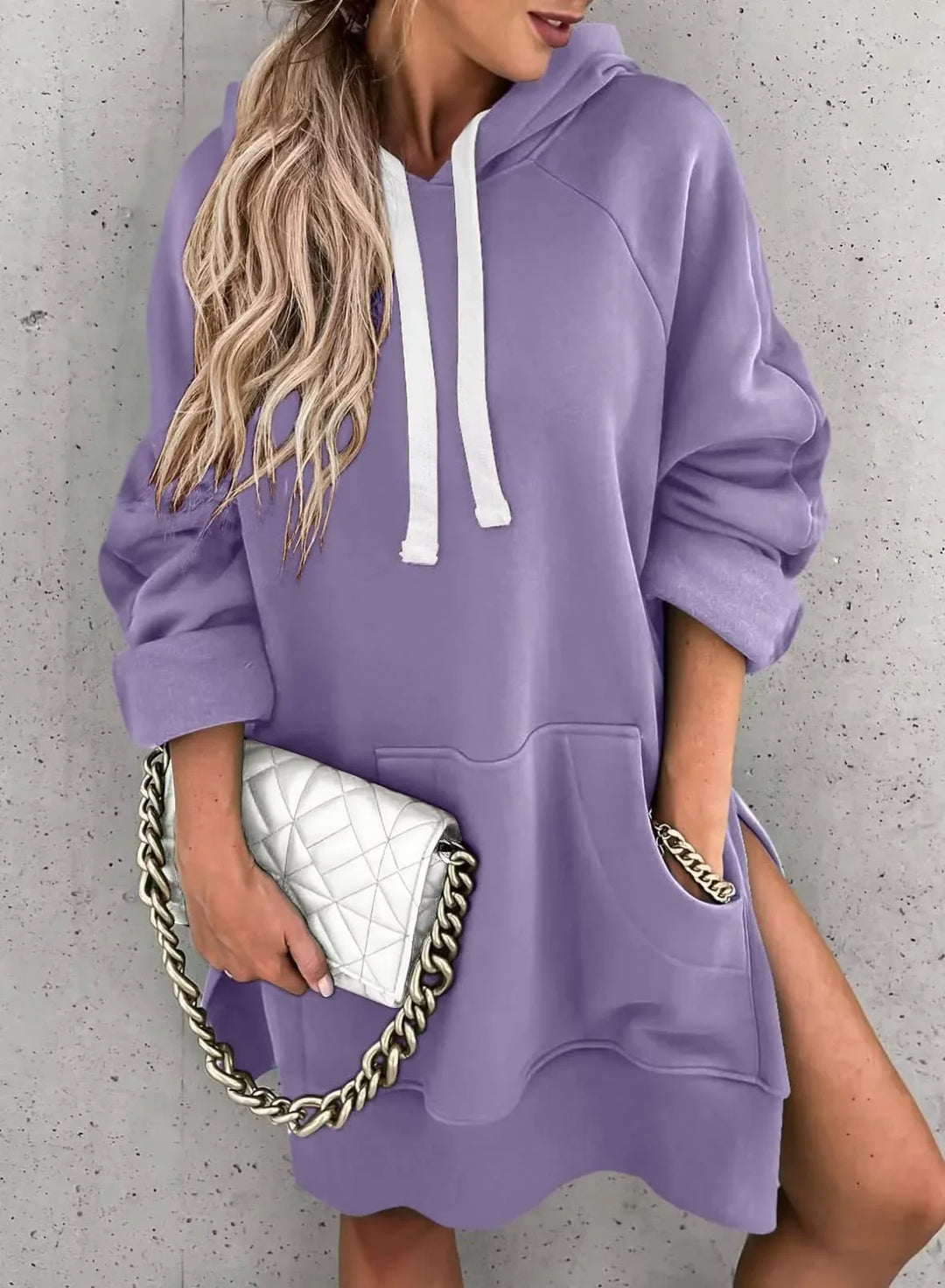 Woman wearing a lilac oversized hoodie dress with white drawstrings, holding a white quilted clutch with a gold chain