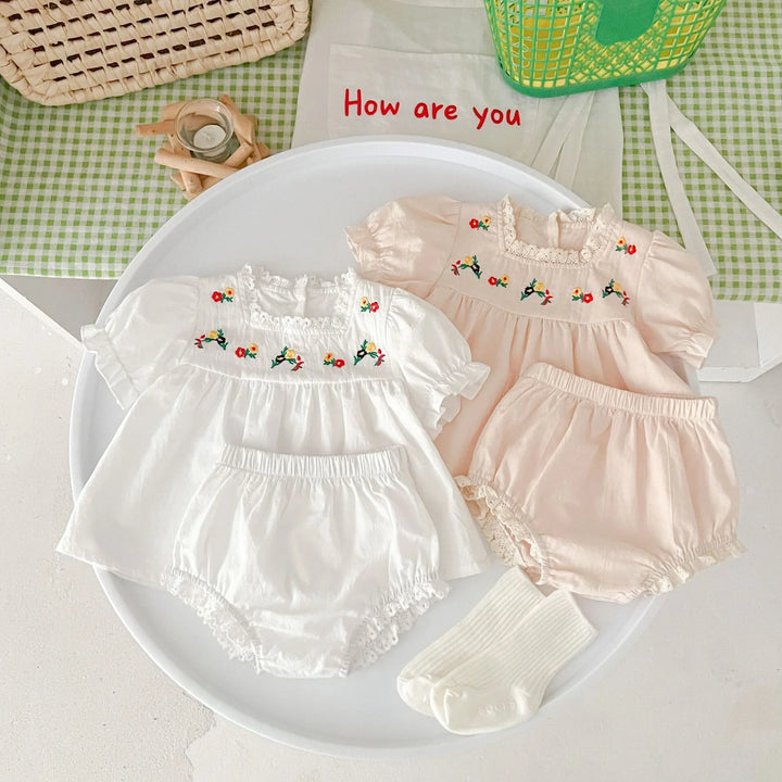 Two baby girl outfits with floral embroidery, one white and one peach, on a round white tray with white socks