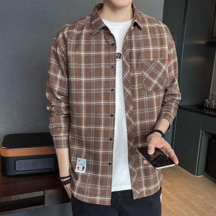Man wearing brown plaid button-up shirt over white t-shirt, holding smartphone indoors
