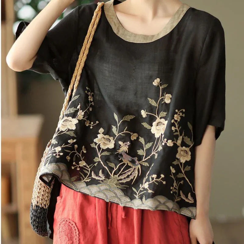 Black linen blouse with beige floral and bird embroidery paired with red pants and a woven shoulder bag