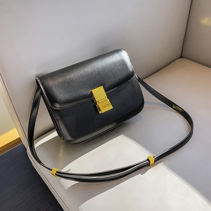 Black leather crossbody bag with gold clasp and adjustable strap on light gray sofa