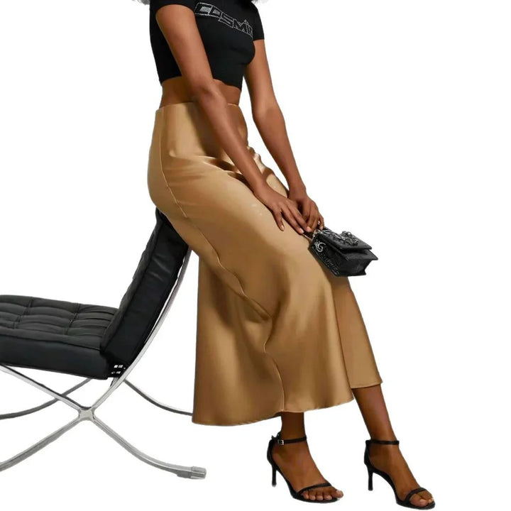 Woman in a tan satin midi skirt, black crop top, black high-heeled sandals, holding a black handbag sitting on modern black leather chair