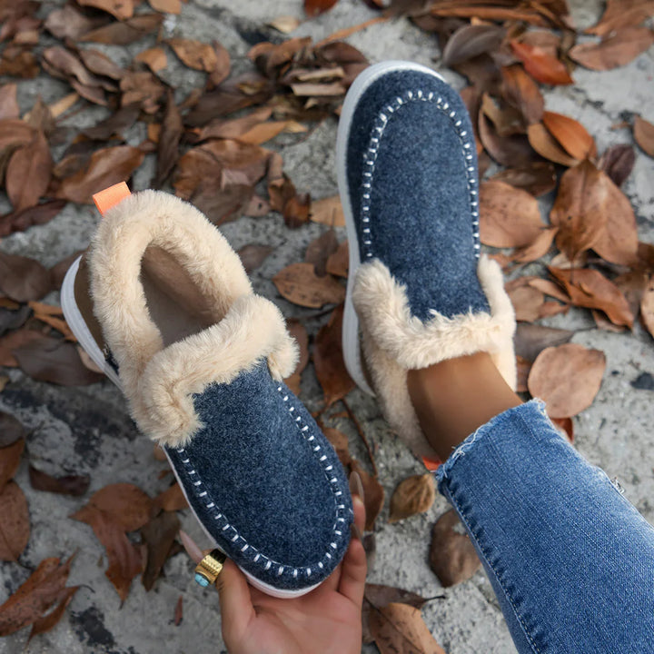 Blue fleece-lined slip-on shoes with white stitching worn outdoors on autumn leaves