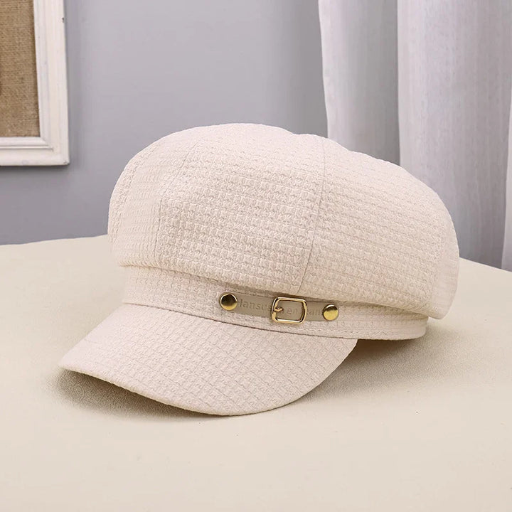 Cream textured newsboy cap with gold buckle detail on a beige surface near white curtain