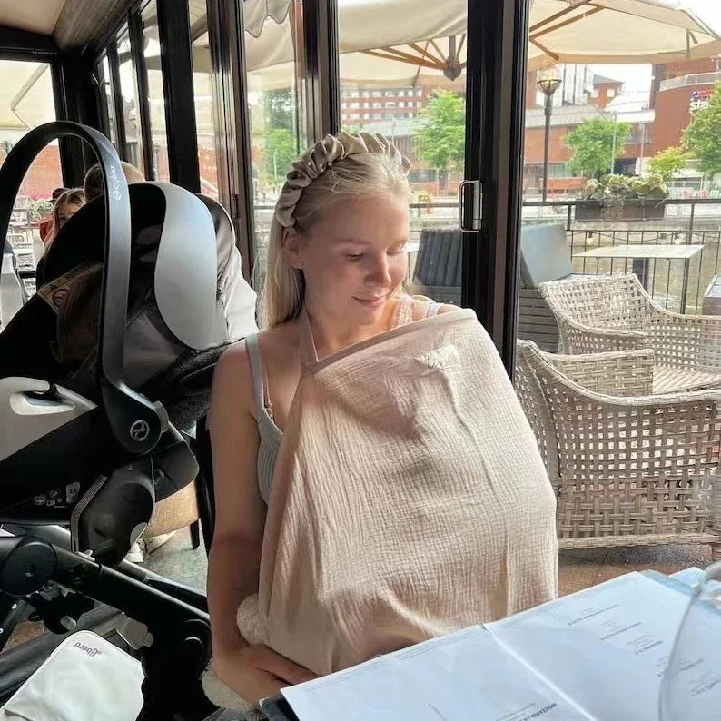 Young woman sitting at a cafe breast-feeding baby under a beige nursing cover with a stroller nearby