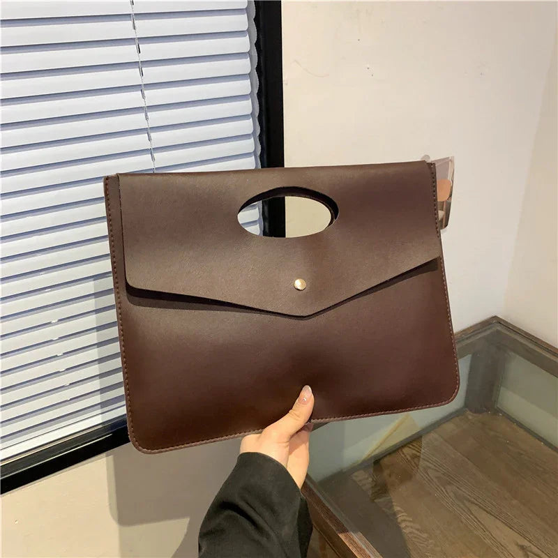 hand holding a sleek brown leather envelope clutch bag with oval handle in a modern indoor setting