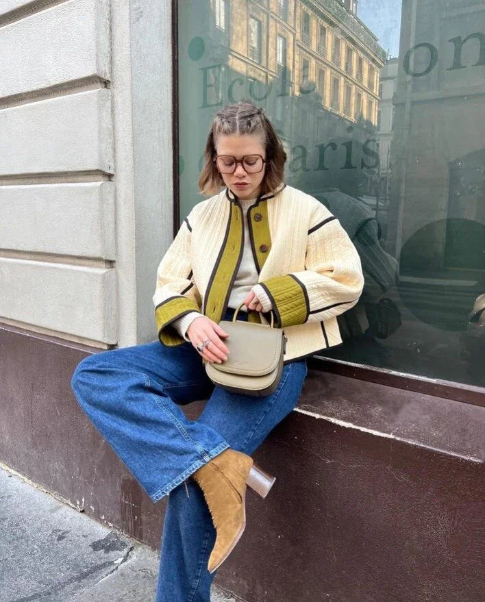 woman wearing cream and green jacket, blue jeans, and tan boots, sitting with beige handbag in urban setting