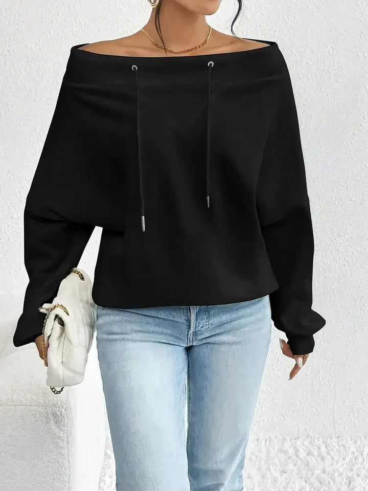 Woman wearing black off-shoulder sweatshirt with drawstrings and light blue jeans holding white handbag