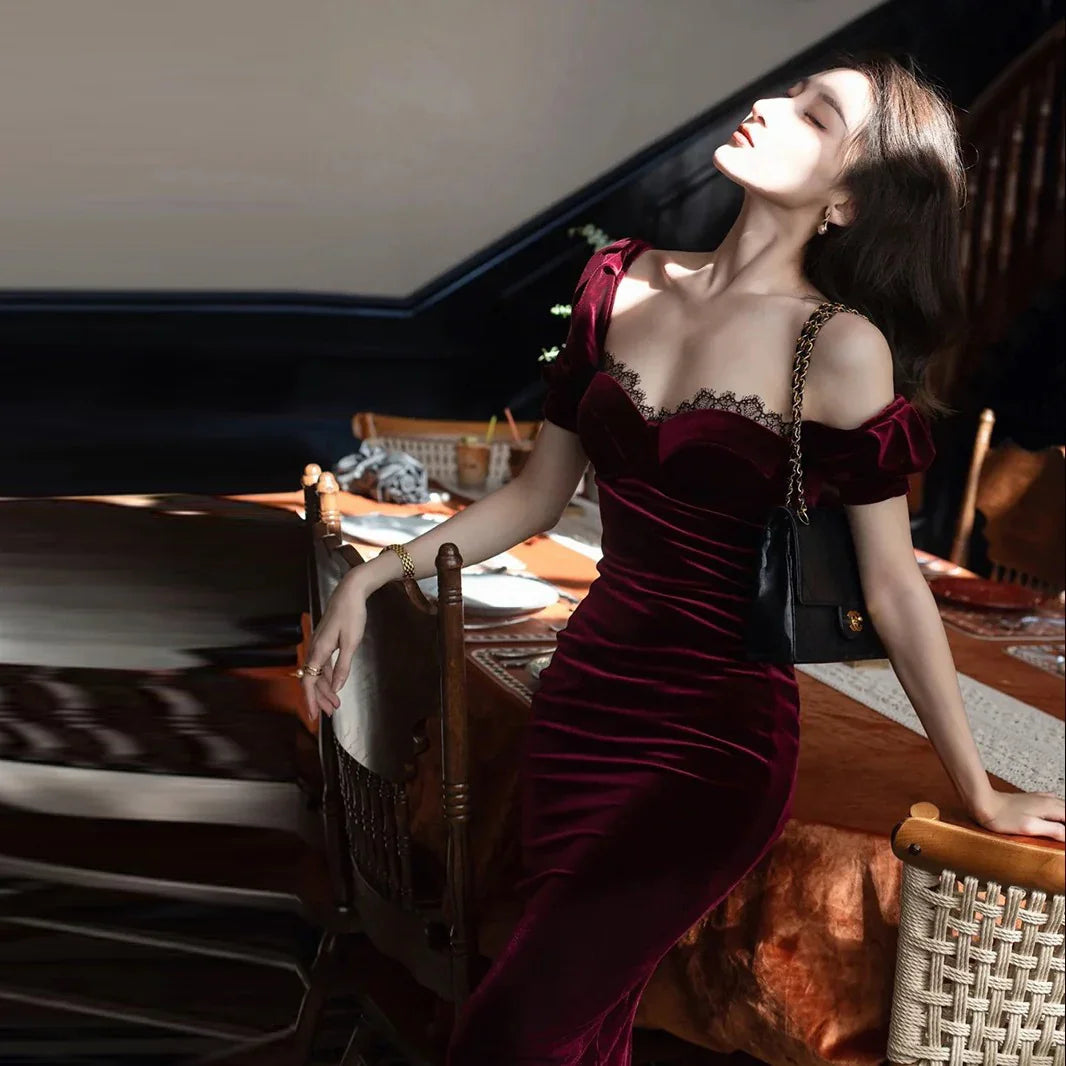 Woman in elegant burgundy velvet dress with lace trim, posing by dining table in soft sunlight