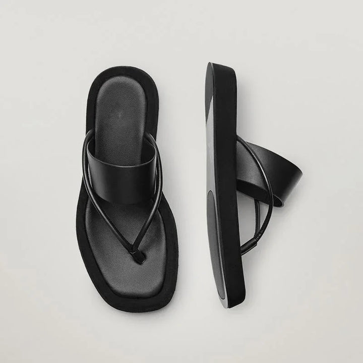 Pair of black minimalist flat sandals with wide strap and thin toe loop on light background