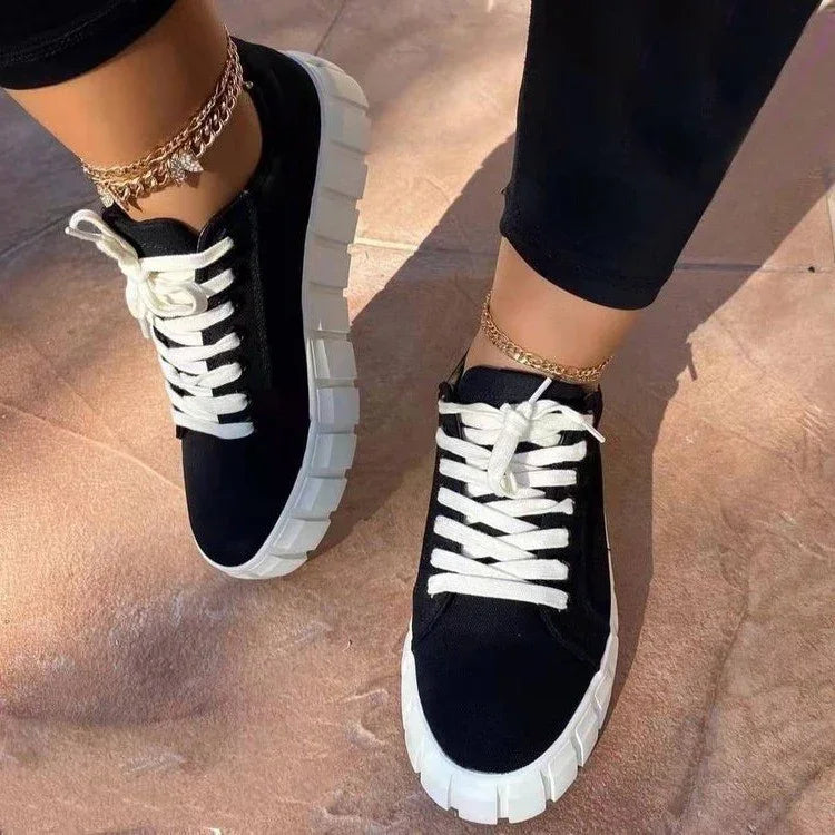 Black canvas sneakers with thick white soles and white laces worn with gold anklets on tiled floor