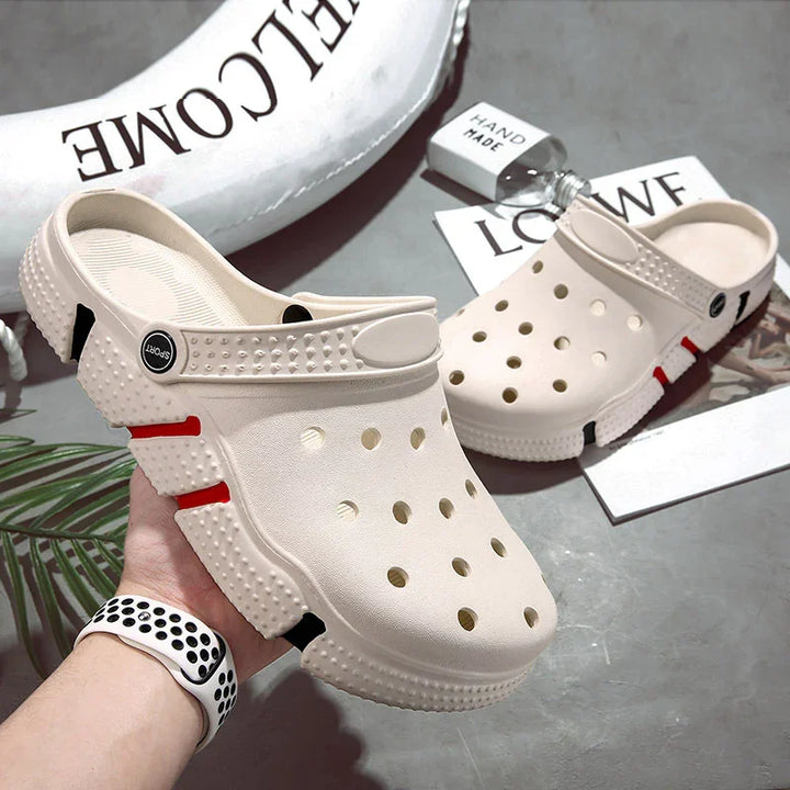 Pair of beige foam clogs with ventilation holes and red-black sole accents on gray surface