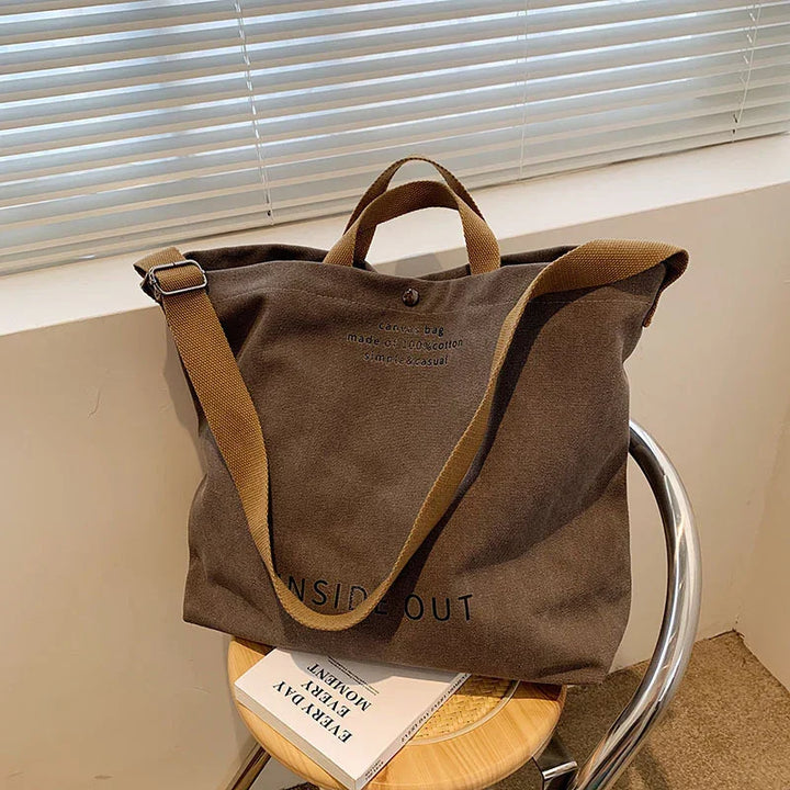 brown canvas shoulder bag with text, placed on wooden stool next to window with blinds