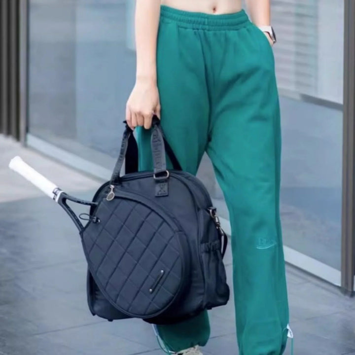 Woman in green sweatpants holding black quilted tennis racket bag outdoors