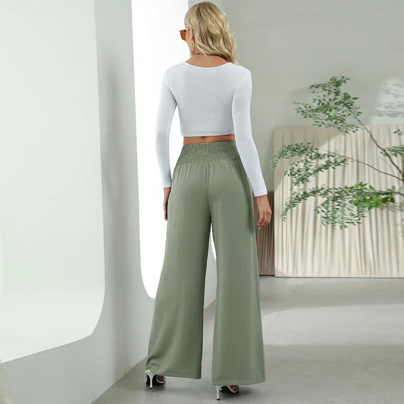 Woman wearing white long-sleeve crop top and sage green wide-leg pants in minimalist room with plant
