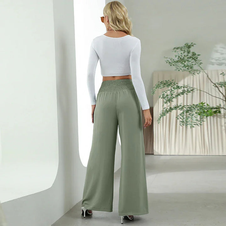 Woman wearing white long-sleeve crop top and sage green wide-leg pants in minimalist room with plant