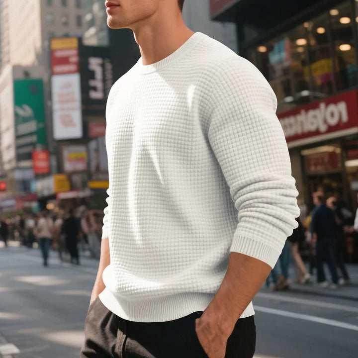 Man wearing white textured knit sweater standing on busy city street with hands in pockets
