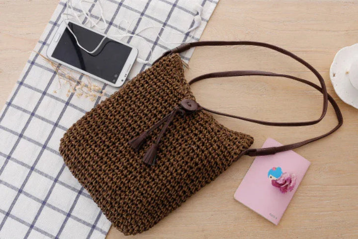 Brown woven crossbody bag with tassel on wooden surface next to smartphone and pink notebook