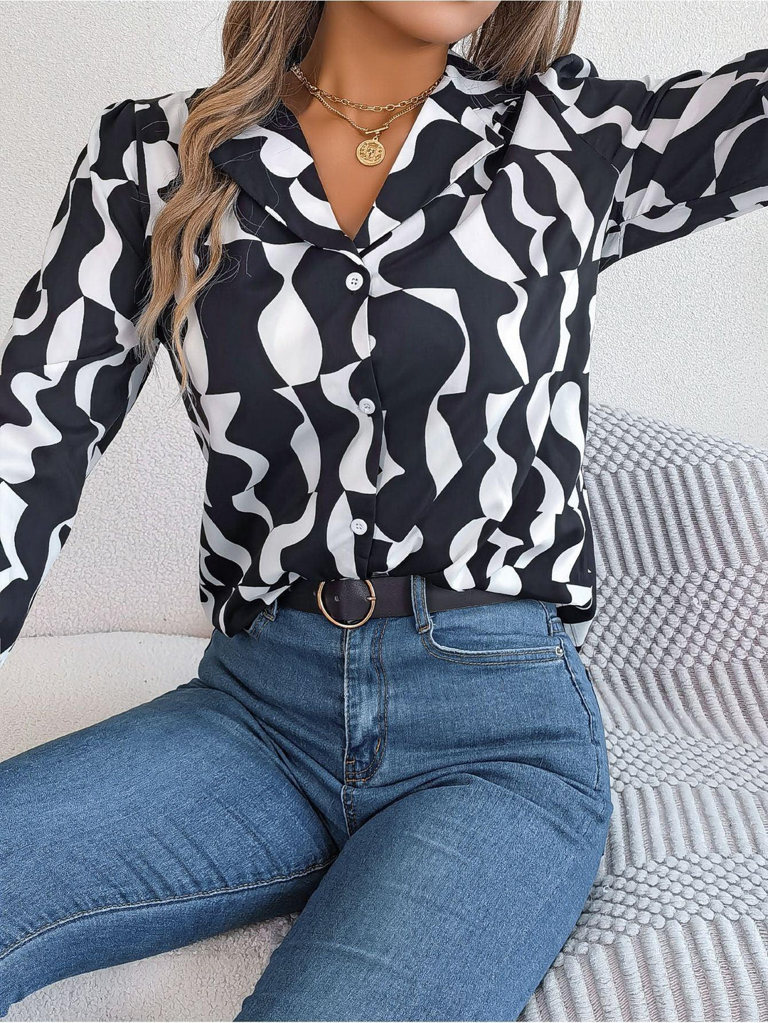 Woman wearing black and white abstract print button-up blouse with blue jeans and gold necklace sitting on textured cushion