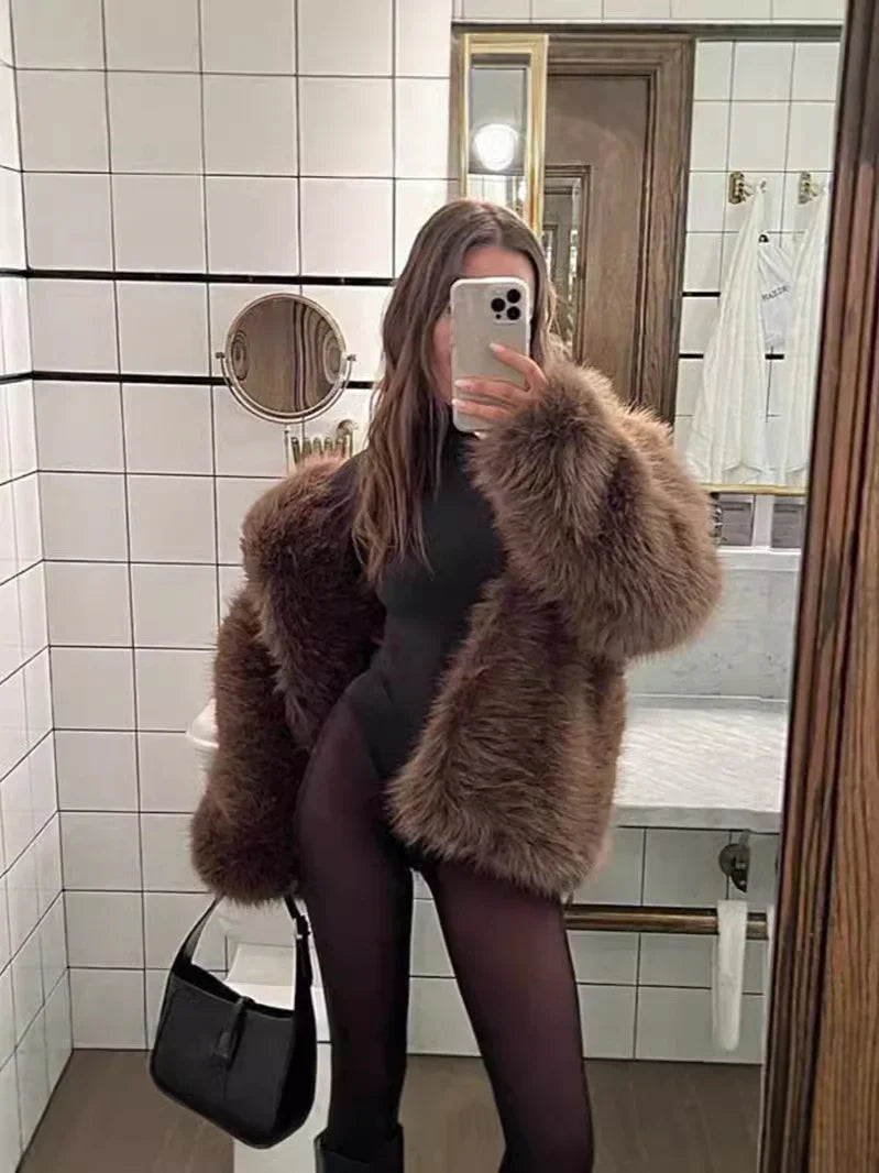 Woman in brown faux fur coat and black outfit taking mirror selfie in modern tiled bathroom