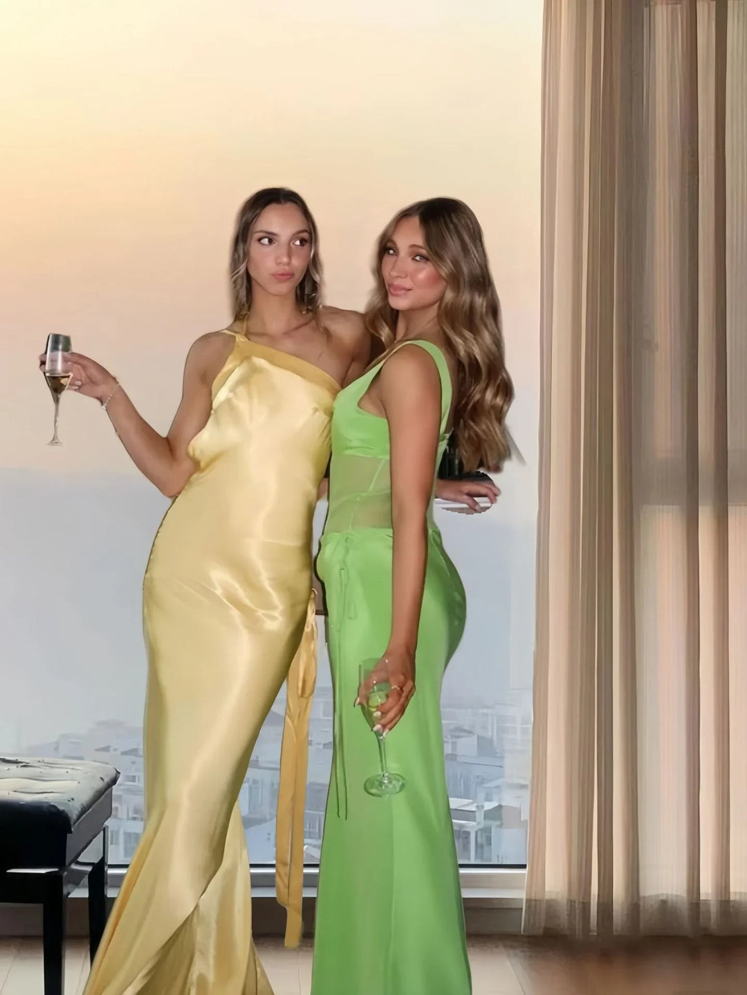 Two women in elegant yellow and green satin dresses holding wine glasses indoors by window