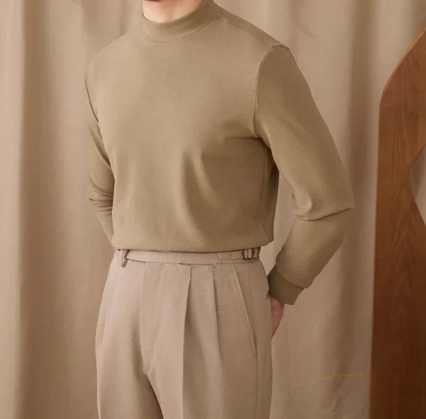 Man wearing beige mock neck sweater and pleated beige trousers against a beige curtain background