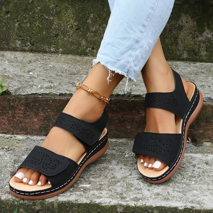 Women's black casual sandals with perforated design, worn with light blue frayed jeans on stone steps