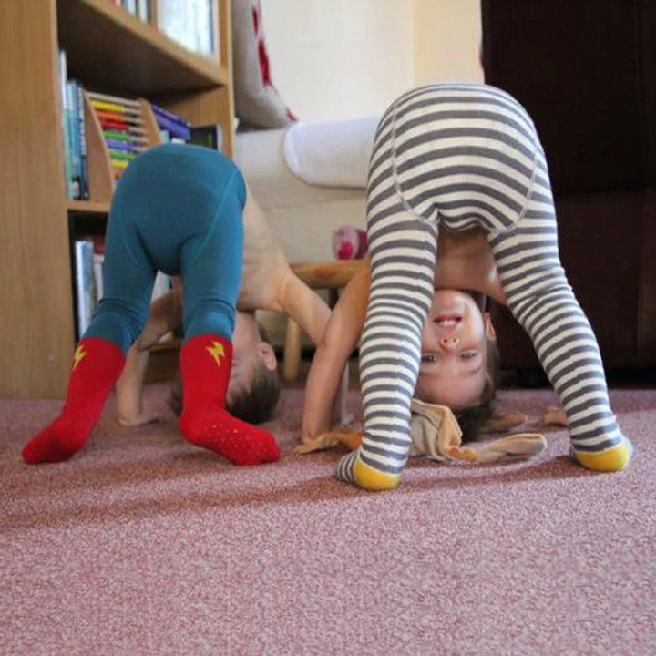 Two children playing indoors, one in blue pants with red socks, the other in gray striped leggings, head down and smiling