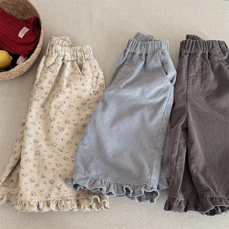 Three pairs of corduroy baby pants with ruffle hems in beige floral, light blue, and brown on beige surface