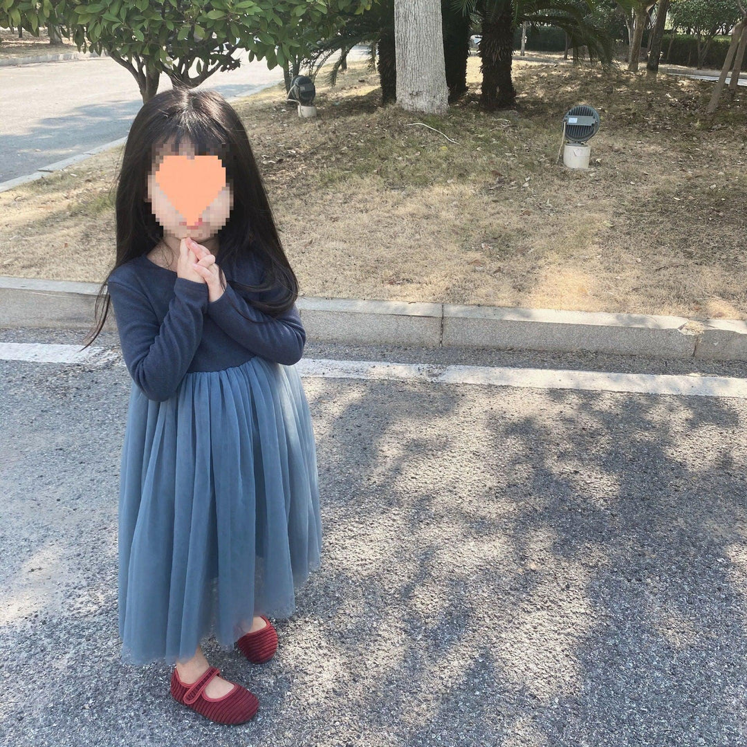 Little girl in long blue dress and red shoes standing on paved path near grass and trees