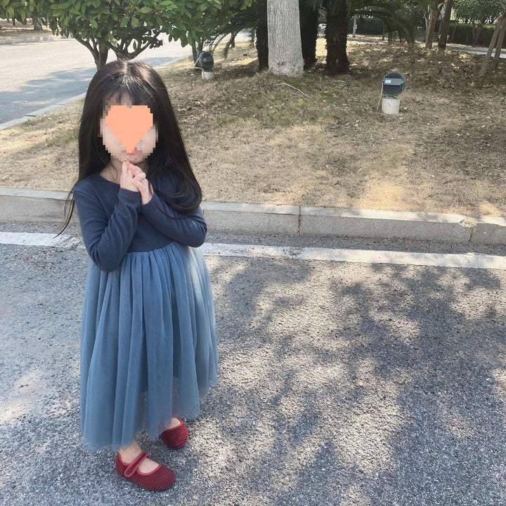 Little girl in long blue dress and red shoes standing on paved path near grass and trees