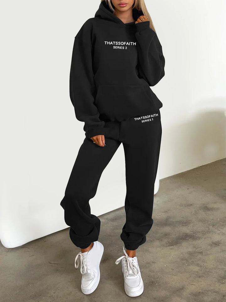 Woman wearing black Thatssofaith Series 2 hoodie and sweatpants with white sneakers on concrete floor