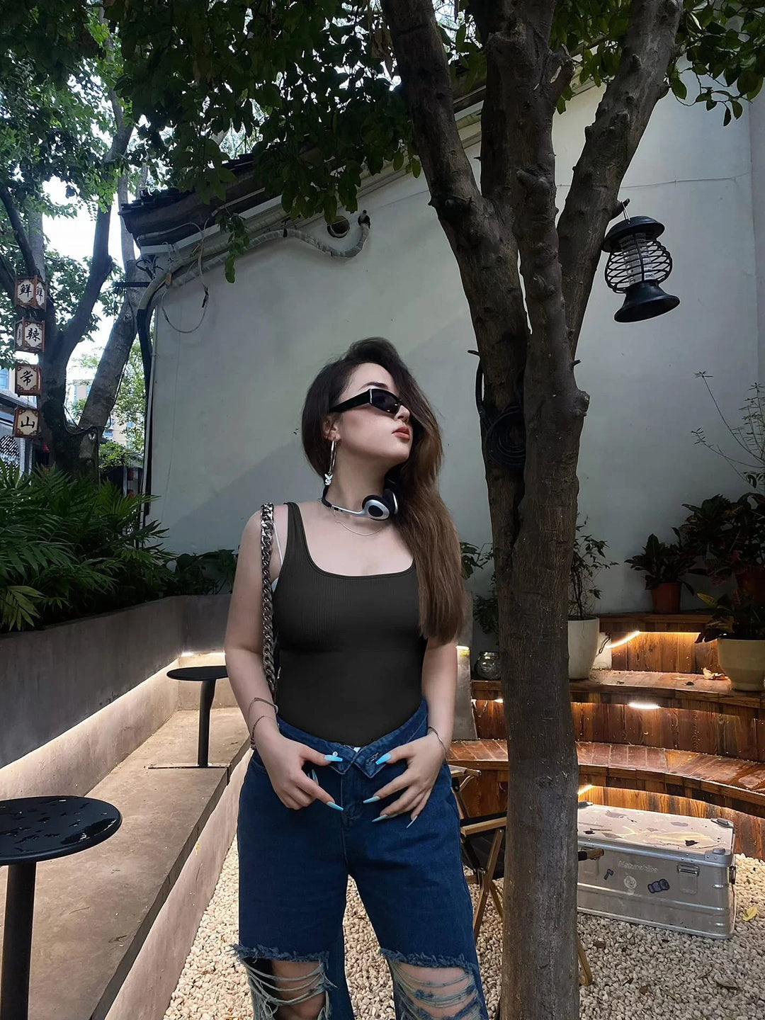Stylish woman with long hair wearing black tank top, ripped blue jeans, sunglasses, and headphones outdoors