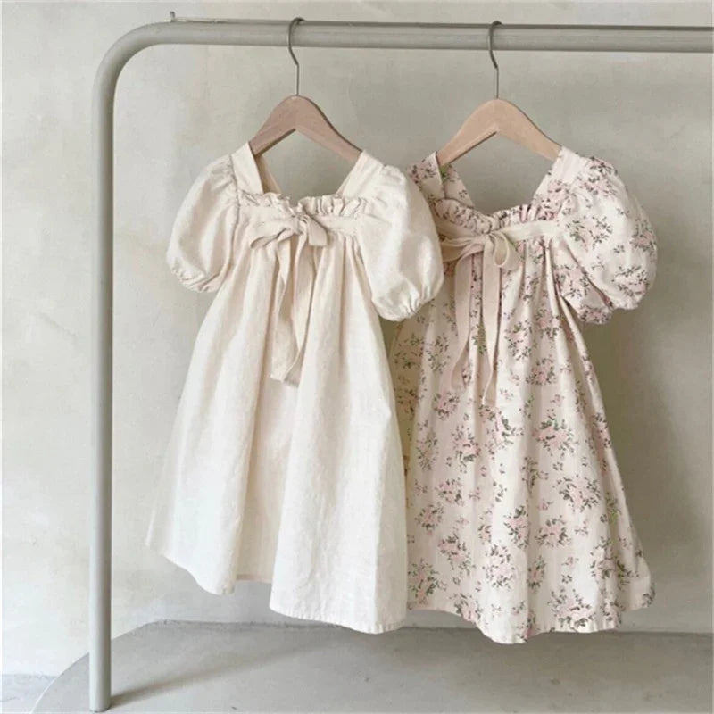 Two children's dresses with puff sleeves hanging on a rack, one plain white and one floral print