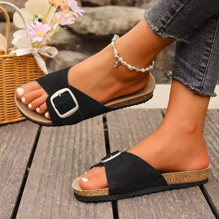 Feet wearing black open-toe slide sandals with silver buckle and beaded ankle bracelet on wooden floor